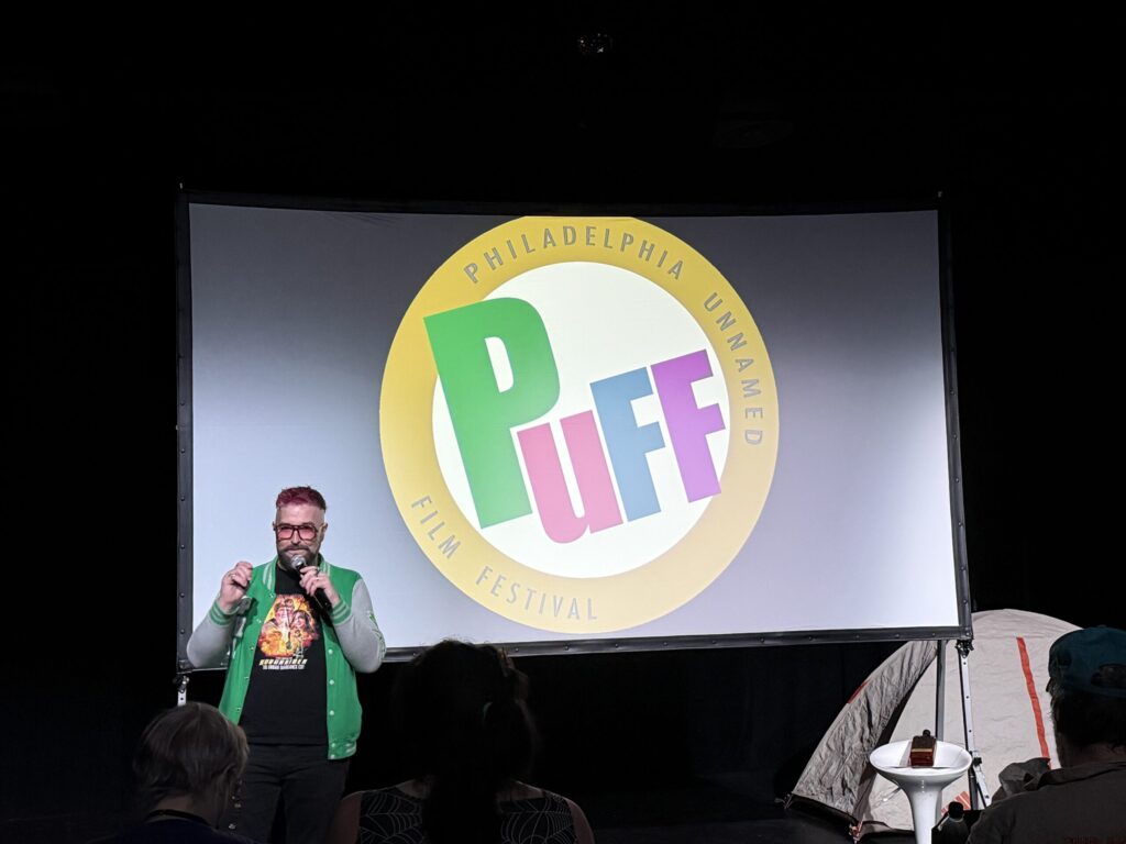 Host Jack Stallion stands in front of a small movie screen that displays the Philadelphia Unnamed Film Festival logo, with a green 'P', red "u", blue "f", and purple "f" in he middle of the circular logo.
