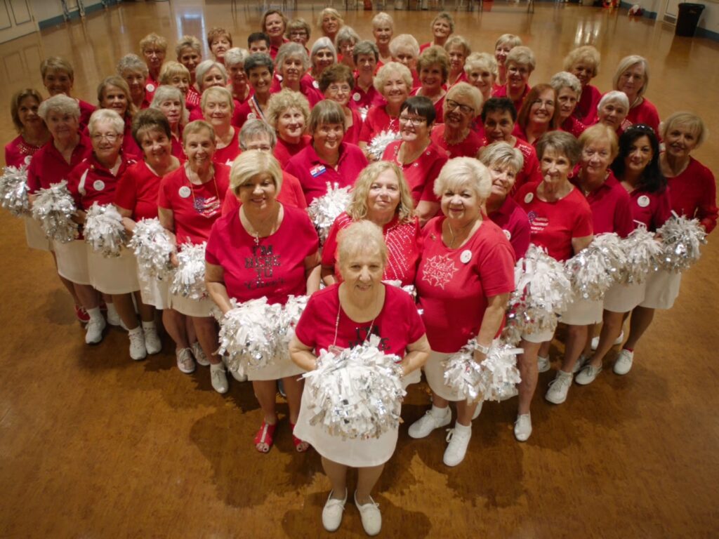A large group of women in  red shirts, white skirts, and white sneakers pose in a hall holding silver/white pom-poms.